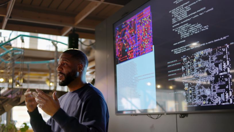 A person presenting in front of a large screen displaying technical content