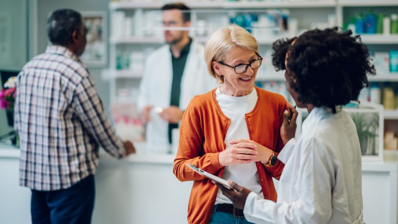 Young black woman pharmacist advising senior female customer about medications in a pharmacy. Shelves with health care products and a male colleague pharmacist in the background.