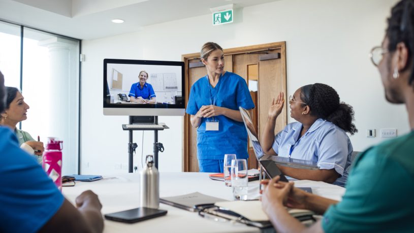 A group of healthcare professionals sitting around a table, taking part in a medical training meeting in a medical building in Whitley Bay, North East England. They are all listening to a webinar on a computer that a nurse is hosting while they listen to another healthcare professional taking questions from the whole group as she stands next to the screen.